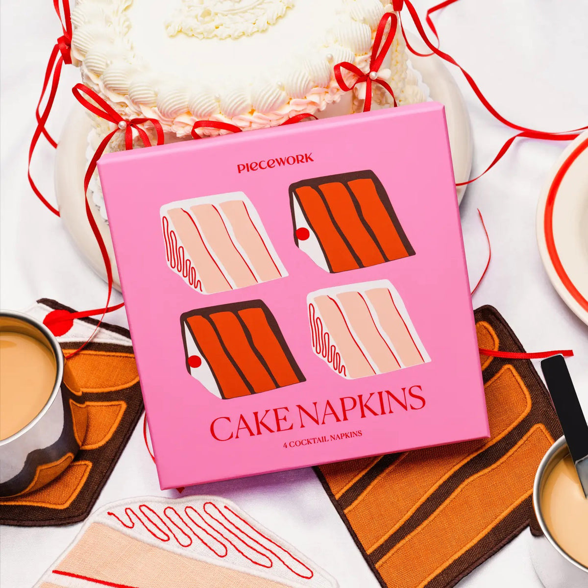 The LINEN CAKE COCKTAIL NAPKINS by PIECEWORK PUZZLES feature four embroidered cotton slices of classic layer cake, complete with cherry and icing details, resting alongside two coffee mugs and red ribbons.