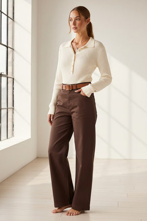 A woman stands barefoot indoors next to a large sunlit window, wearing the MERSEA - LUDLOW LONG SLEEVE POLO IN CREME with a collared neckline, paired with high-waisted brown wide-leg pants and a belt.