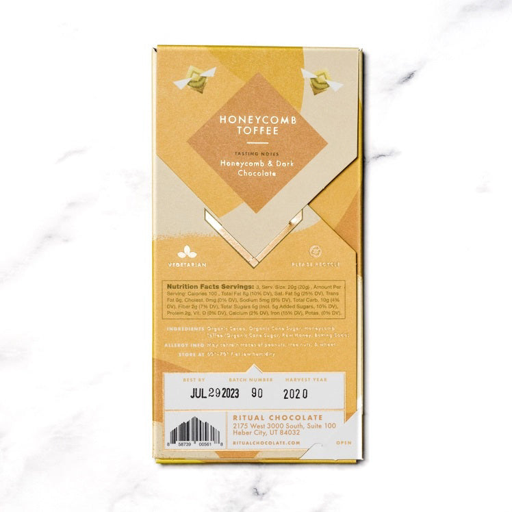 A gold and yellow RITUAL CHOCOLATE - HONEYCOMB TOFFEE CHOCOLATE BAR wrapper with geometric designs holds a 2.12 oz (60 g), 75% cacao single origin dark chocolate bar, displayed on a marble background.