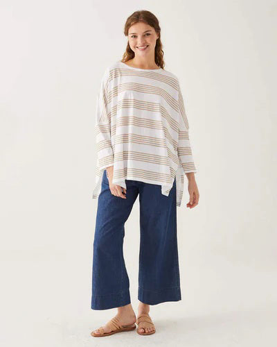 A woman with wavy brown hair models the MERSEA - CATALINA SLUB TEE—an oversized, long-sleeved white top with thin gray stripes—by MERSEA. She pairs it with blue jeans and stands against a plain, light background, hands behind her back.
