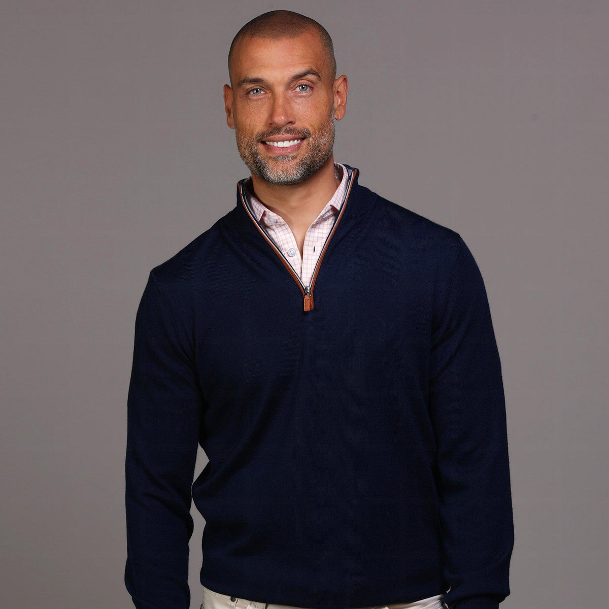 A man with short hair and a beard stands against a gray background wearing the COLLARS AND CO Navy with Brown Suede Trim Cashmere Touch Australian Merino Quarter Zip Sweater over a white shirt, khaki pants, and brown high-top sneakers, one hand in his pocket.
