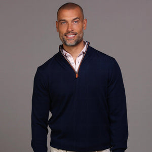 A man with short hair and a beard stands against a gray background wearing the COLLARS AND CO Navy with Brown Suede Trim Cashmere Touch Australian Merino Quarter Zip Sweater over a white shirt, khaki pants, and brown high-top sneakers, one hand in his pocket.