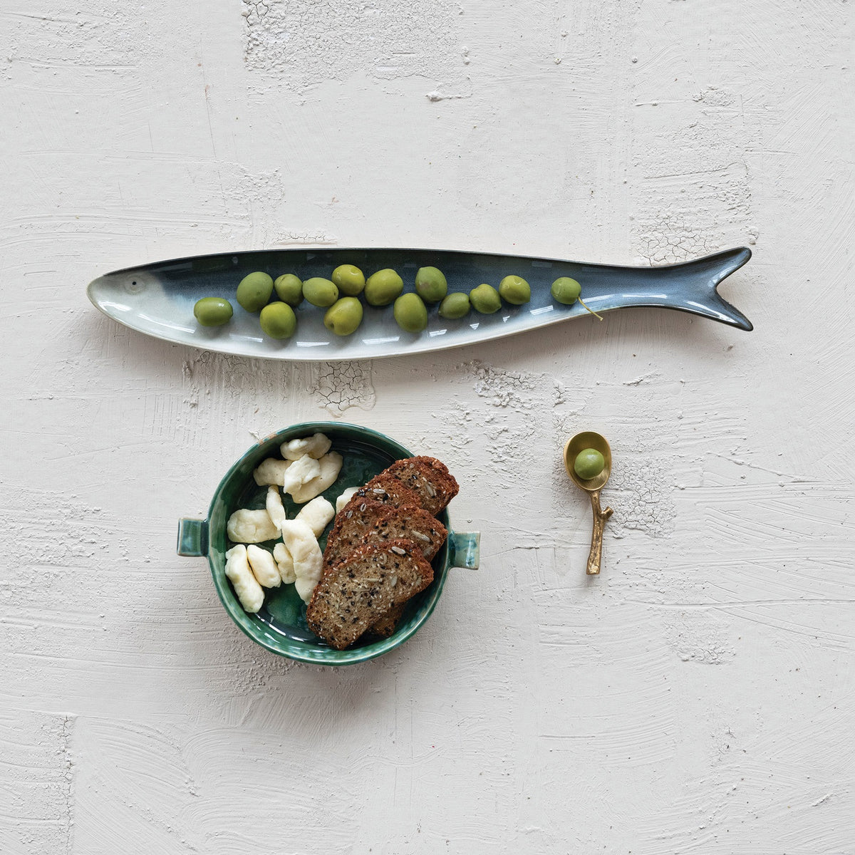 A fish-shaped serveware dish with green olives, a small bowl of bread and cheese pieces, and the CREATIVE COOP BRASS SPOON WITH TWIG SHAPED HANDLE holding an olive, all displayed on a textured white surface.