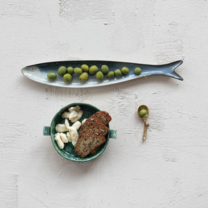 A fish-shaped serveware dish with green olives, a small bowl of bread and cheese pieces, and the CREATIVE COOP BRASS SPOON WITH TWIG SHAPED HANDLE holding an olive, all displayed on a textured white surface.