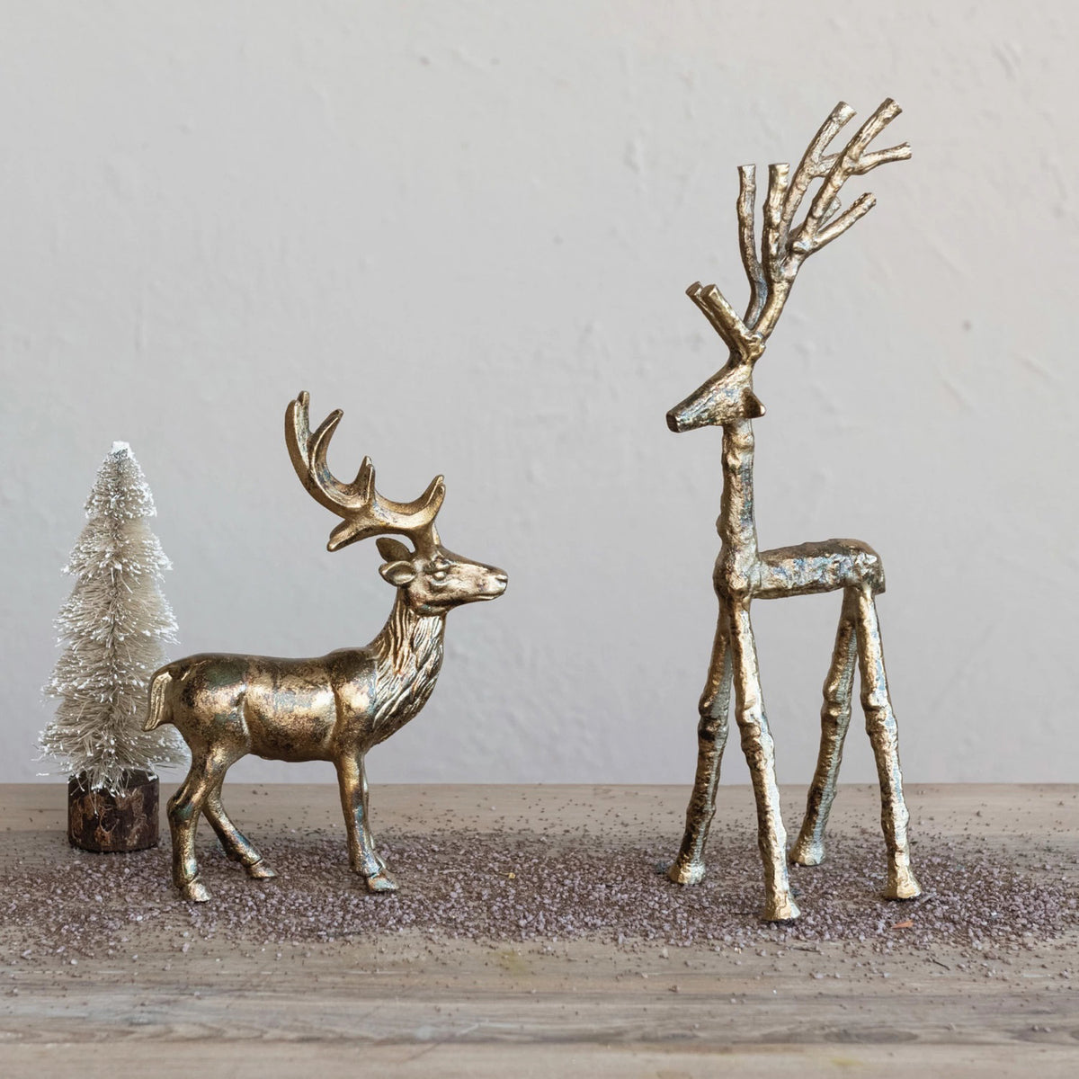 Two metallic reindeer figurines—a CREATIVE COOP CAST ALUMINUM TWIG DEER in an antique gold finish and an abstract long-legged deer—stand beside a small white bottlebrush tree on a glitter-dusted wooden surface.