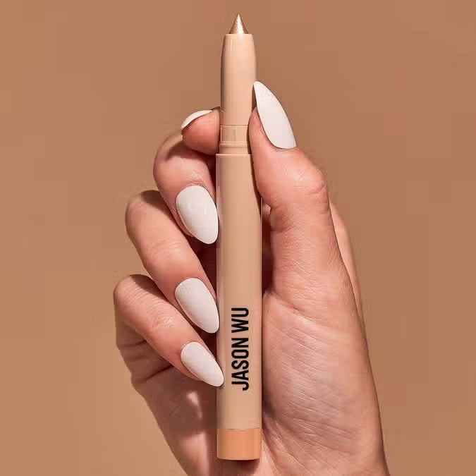 Close-up of a hand with white painted nails holding a sparkle pearl colored eyeshadow pencil with a pink tip, branded with &quot;JASON WU BEAUTY&quot; in black text against a solid neutral beige background.