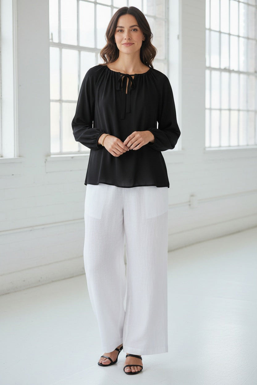 A woman in the MEIMEIJ Tie Neck Blouse—loose, long-sleeved, peach-colored washable silk with a neckline tie—paired with white pants, stands against a light textured background. Her face is partially out of frame.