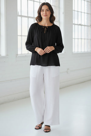 A woman in the MEIMEIJ Tie Neck Blouse—loose, long-sleeved, peach-colored washable silk with a neckline tie—paired with white pants, stands against a light textured background. Her face is partially out of frame.