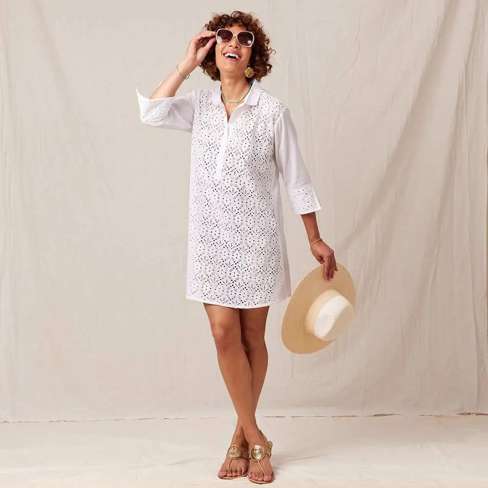 A woman wears the ROCKFLOWERPAPER WHITE EYELET BEACH COVERUP, styled with sunglasses, sandals, and a straw hat, standing against a neutral fabric backdrop.
