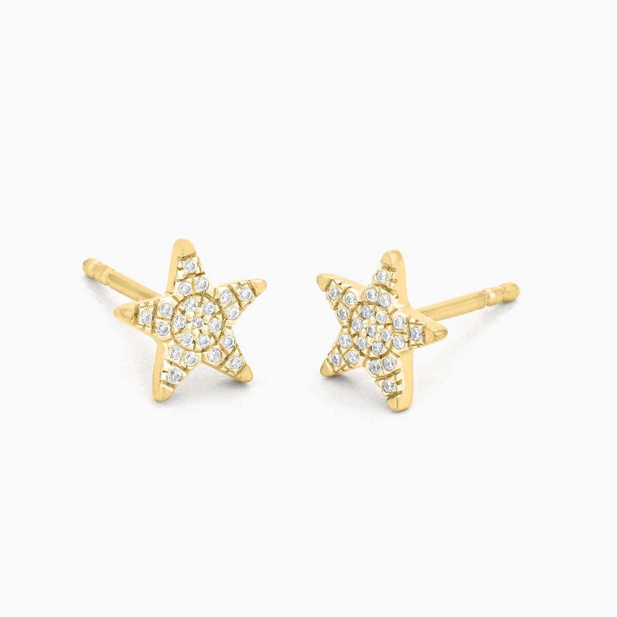 A close-up of a woman’s ear showcases the ELLA STEIN - REACH FOR THE STARS STUD EARRINGS by ELLA STEIN, delicately gold plated and shaped like a tiny star. The softly blurred background features hints of green leaves.