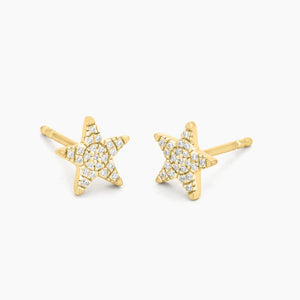 A close-up of a woman’s ear showcases the ELLA STEIN - REACH FOR THE STARS STUD EARRINGS by ELLA STEIN, delicately gold plated and shaped like a tiny star. The softly blurred background features hints of green leaves.
