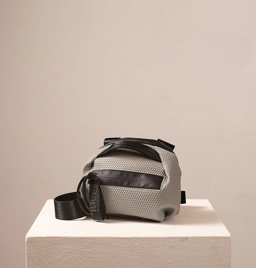 The DANIELLA LEHAVI BONNIE MINI TRAVEL CROSSBODY, a small brown vegan bag with black accents and strap, is displayed on a white pedestal against a plain beige background.