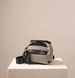 The DANIELLA LEHAVI BONNIE MINI TRAVEL CROSSBODY, a small brown vegan bag with black accents and strap, is displayed on a white pedestal against a plain beige background.