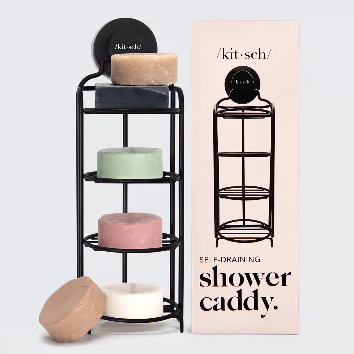 The image depicts a black, rust-proof SELF DRAINING SHOWER CADDY by KITSCH holding four round shampoo bars in various colors: pink, green, white, and grey. Next to the eco-friendly caddy is a tall box featuring an illustration of the caddy and labeled with "/kit·sch/ SELF DRAINING SHOWER CADDY.