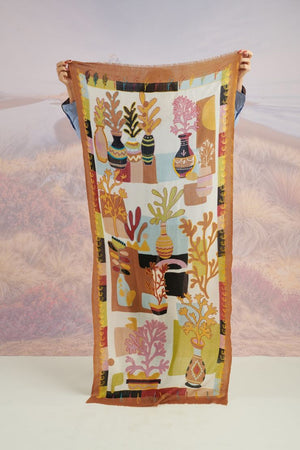 A woman with light brown hair wears a cream blouse and the STORIATIPIC - ANDRES WOOL SCARF by STORIATIPIC, elegantly hand-embroidered with green, yellow, red, and blue patterns. She gazes to the side against a scenic background.