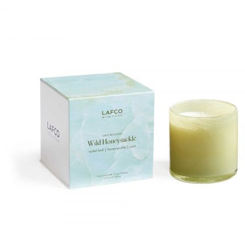 A LAFCO - Wild Honeysuckle Signature 6.5oz candle sits next to its matching box. The box, labeled &quot;Wild Honeysuckle,&quot; lists the scent notes as &quot;violet leaf, honeysuckle, white.&quot; This vegan candle is housed in a light green, hand-blown glass vessel. Both items are elegantly placed against a white background.
