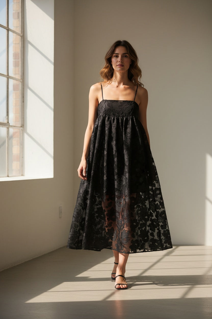 A woman wears the MEIMEIJ Lace Empire Waist Dress, a sleeveless black floral lace midi with spaghetti straps and a sheer overlay. The flowing fit makes it perfect for a garden party. She stands against a plain white background.