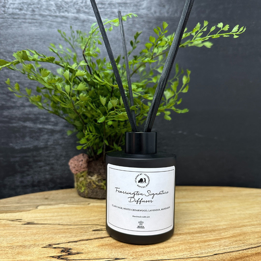 A matte black glass bottle containing several thin black reeds makes an ideal holiday gift. The label on the front reads &quot;Fearrington Signature Diffuser - Clary Sage, White Cedarwood, Calming Lavender, Marjoram.&quot; The backdrop is a white, textured surface.