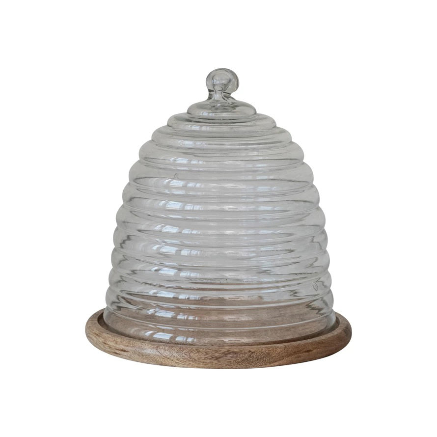 Introducing the BEE HIVE CLOCHE WITH TRAY by CREATIVE COOP: This eco-friendly serveware piece features a glass cloche shaped like a beehive with grooves resembling hive layers and now includes a sleek mango wood base. It comes with a spherical knob on top for easy lifting, and the cloche itself is made from reclaimed glass for added sustainability.