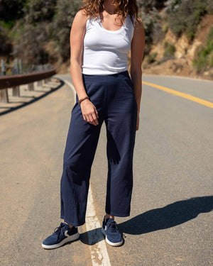 A woman stands in a minimal white room wearing the MERSEA TRAVELER TEE PANT, light gray, made from Turkish cotton slub—styled with white sneakers featuring green accents—and smiles softly at the camera.