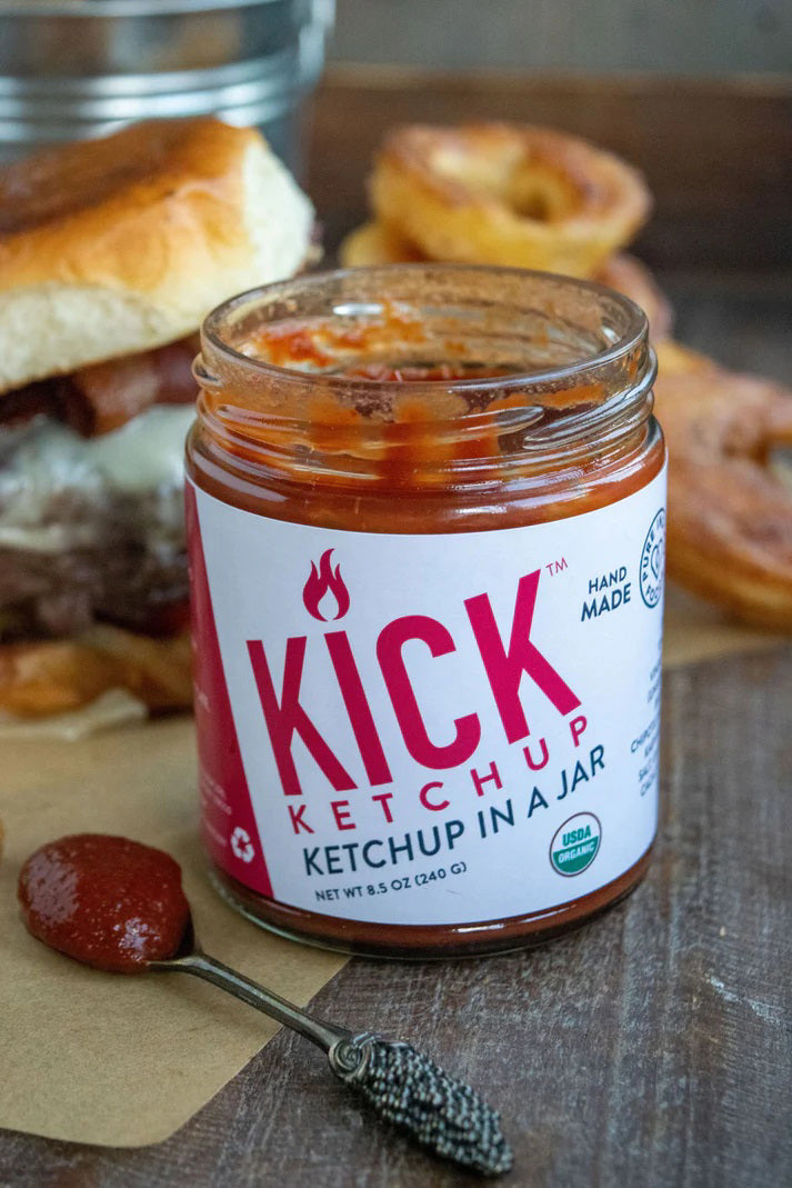 A jar of PURE INDIAN FOODS- KICK KETCHUP by PURE INDIAN FOODS features a black lid, white label with bold red text, flame icon, and &quot;Ketchup in a Jar.&quot; This spicy ketchup is handmade with chipotle morita and is USDA Organic.