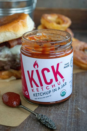 A jar of PURE INDIAN FOODS- KICK KETCHUP by PURE INDIAN FOODS features a black lid, white label with bold red text, flame icon, and "Ketchup in a Jar." This spicy ketchup is handmade with chipotle morita and is USDA Organic.