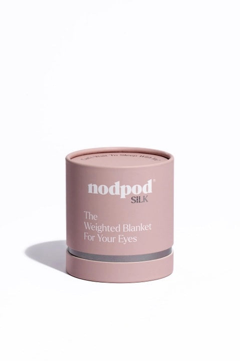 A woman in a gray bralette and shorts lies on white bedding, wearing the NODPOD Silk Weighted Sleep Eye Mask in Petal—a large pink satin silk mask shaped like a bow from NODPOD that covers her eyes for comfort and stress relief.