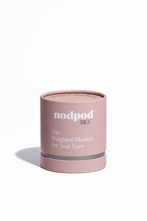 A woman in a gray bralette and shorts lies on white bedding, wearing the NODPOD Silk Weighted Sleep Eye Mask in Petal—a large pink satin silk mask shaped like a bow from NODPOD that covers her eyes for comfort and stress relief.