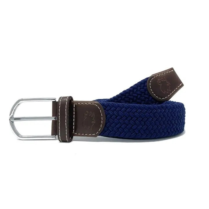 A person outdoors with hands in pockets, wearing a navy blue shirt, light blue pants, and the ROOSTAS PEEBLE BEACH WOVEN STRETCH BELT by ROOSTAS.