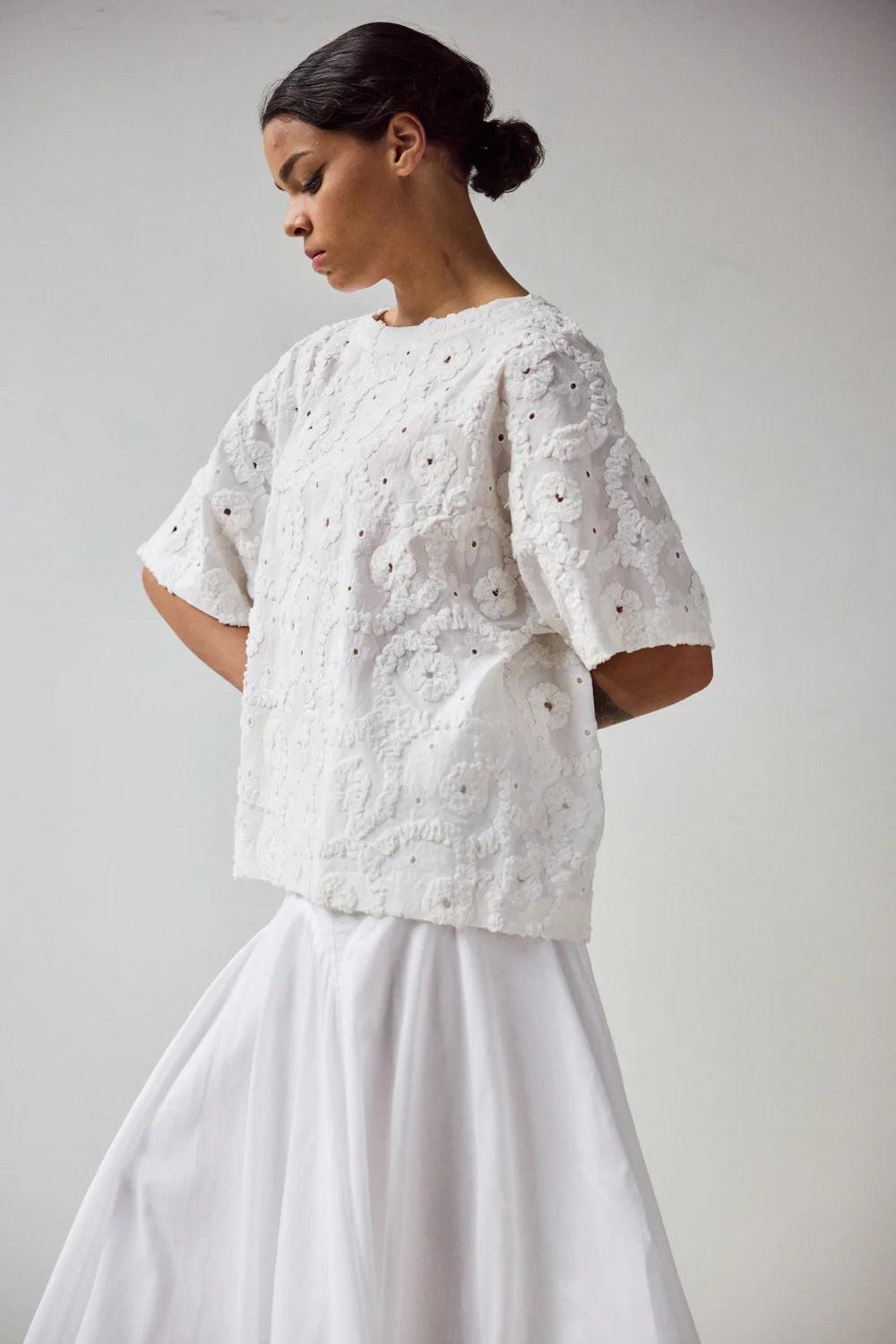 A woman stands in profile wearing the HOPE FOR FLOWERS EYELET TEE, a white, short-sleeved textured lace top made from GOTS certified organic cotton, paired with a flowing white skirt. Her hair is pulled back with a plain light background.