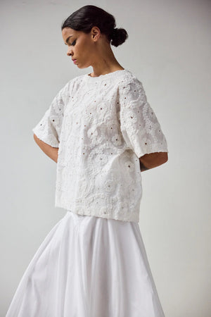 A woman stands in profile wearing the HOPE FOR FLOWERS EYELET TEE, a white, short-sleeved textured lace top made from GOTS certified organic cotton, paired with a flowing white skirt. Her hair is pulled back with a plain light background.