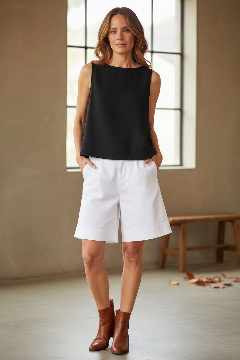 A woman stands outside in a garden with a white building behind her, smiling and looking to the side. She is wearing the FRANK &amp; EILEEN - SHELLEY Shell Tank in Famous Denim Black—a sleeveless, artisan-made top with back buttons—paired with white shorts.
