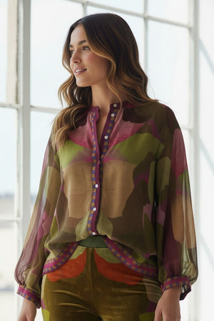 A woman stands indoors, smiling, wearing the FRANCO FERRARI - GERRY SILK TOP featuring an abstract botanical print and button details, paired with blue jeans. Sunlight from a large window illuminates the room, with greenery visible outside.