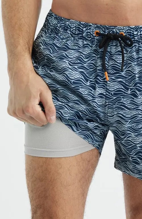 A shirtless man stands with hands in his pockets against a plain light background, modeling SAVE THE DUCK's Ademir Swim Trunks in Ondine Off White on Blue, crafted from quick-dry recycled polyester.