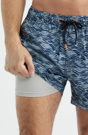 A shirtless man stands with hands in his pockets against a plain light background, modeling SAVE THE DUCK's Ademir Swim Trunks in Ondine Off White on Blue, crafted from quick-dry recycled polyester.