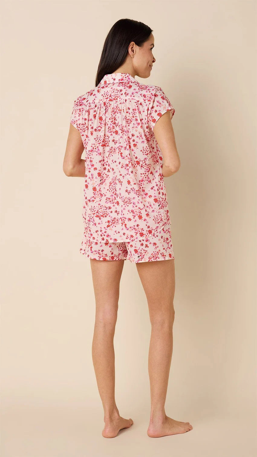 A smiling woman stands barefoot against a beige background, wearing the CAT&#39;S PAJAMAS - PETITE ROSETTE VOILE SHORT SET by CAT&#39;S PAJAMAS—a pink and white floral cotton voile pajama top and matching shorts—ideal for lightweight women&#39;s sleepwear.