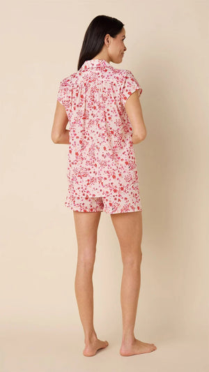 A smiling woman stands barefoot against a beige background, wearing the CAT'S PAJAMAS - PETITE ROSETTE VOILE SHORT SET by CAT'S PAJAMAS—a pink and white floral cotton voile pajama top and matching shorts—ideal for lightweight women's sleepwear.