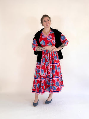 A smiling woman in the FARYLINA - CAMILLE LONG DRESS, a bold blue and pink floral cotton dress by FARYLINA, stands against a white background with one hand on her hip, showing arm tattoos and wearing gray flats.