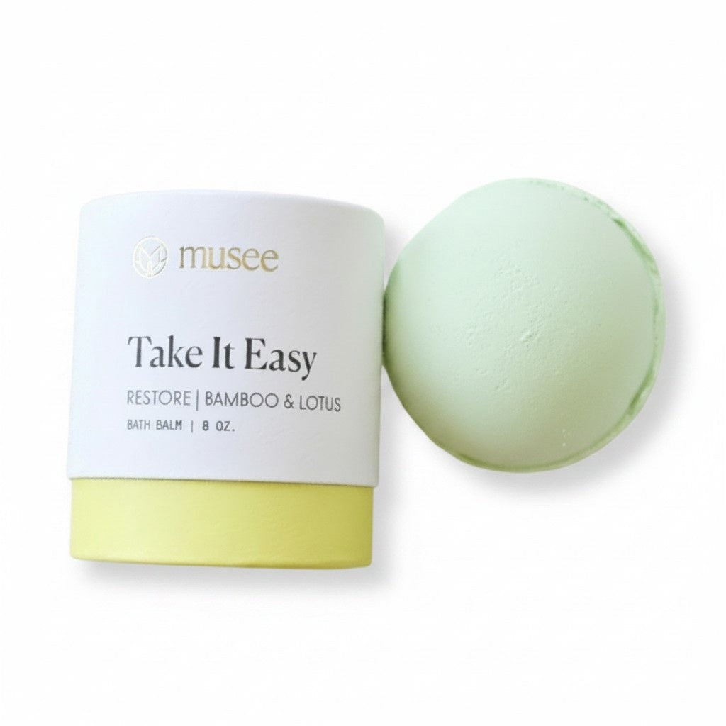 A light green MUSEE - TAKE IT EASY BATH BALM by MUSEE sits on a white plate next to a decorative plant atop a wooden tray, highlighting the artisanal appeal of handcrafted bath products.