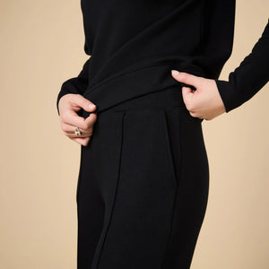 A woman wears a SOFTIES - DREAMTECH BOAT NECK WITH PINTUCK PANT SET in olive green, featuring a long-sleeve boat neck top and matching wide-leg pintuck pants.
