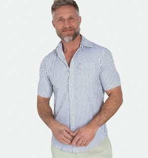 A man with a gray beard wears the CRIQUET Short Sleeve Performance Seersucker Pearl Snap Shirt in Navy while standing on a boat, with water, palm trees, and cloudy skies in the background.