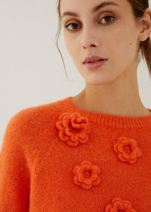A woman with light brown hair wears the ANNECLAIRE hand embroidered crew neck wool sweater in light green with flower details and a white skirt, standing against a pale background. The timeless piece is handmade in Italy.