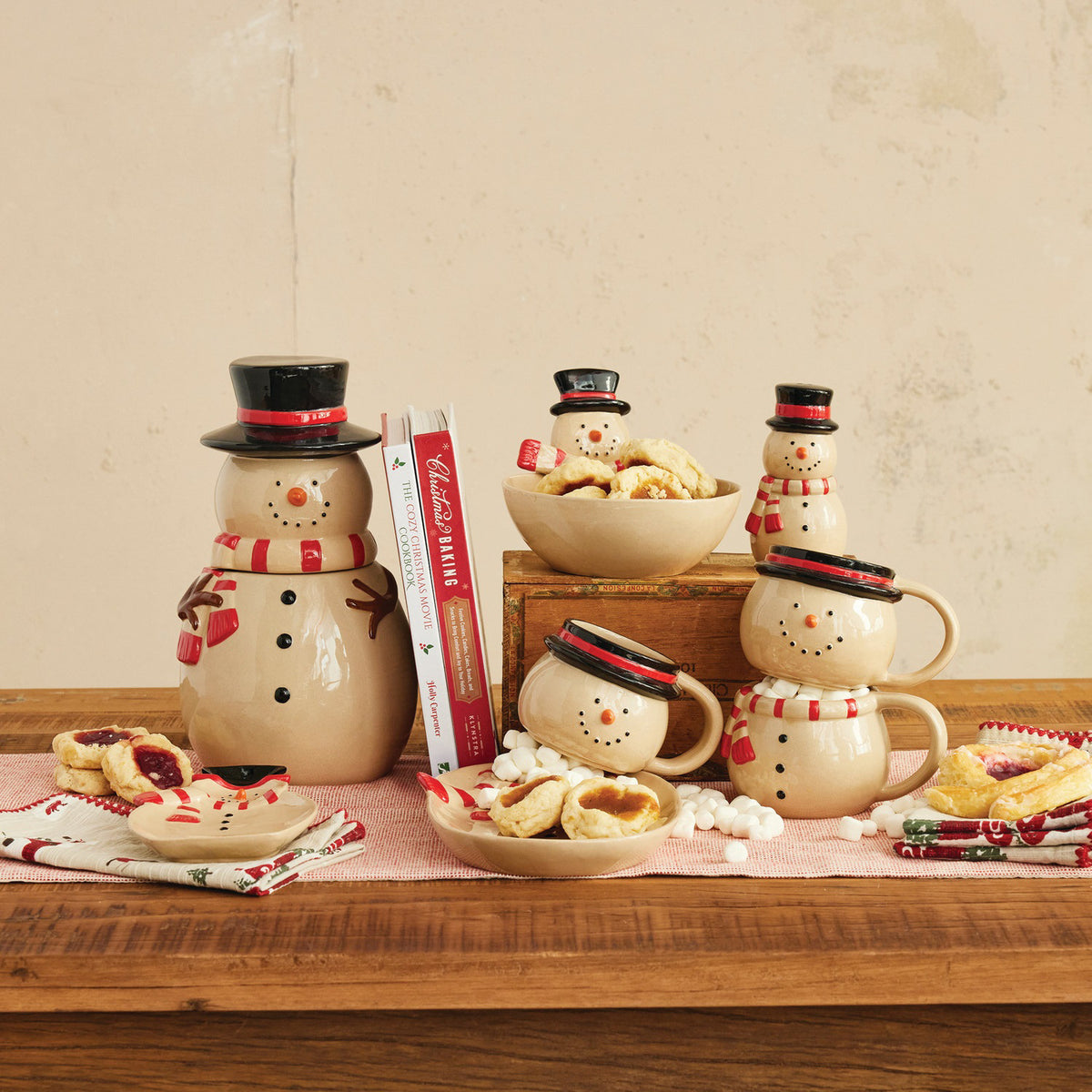 A cozy holiday table features the CREATIVE COOP STONEWARE BOWL WITH SNOWMAN HEAD, filled with treats and surrounded by snowman mugs and a teapot, all on a wooden table with a pink cloth.
