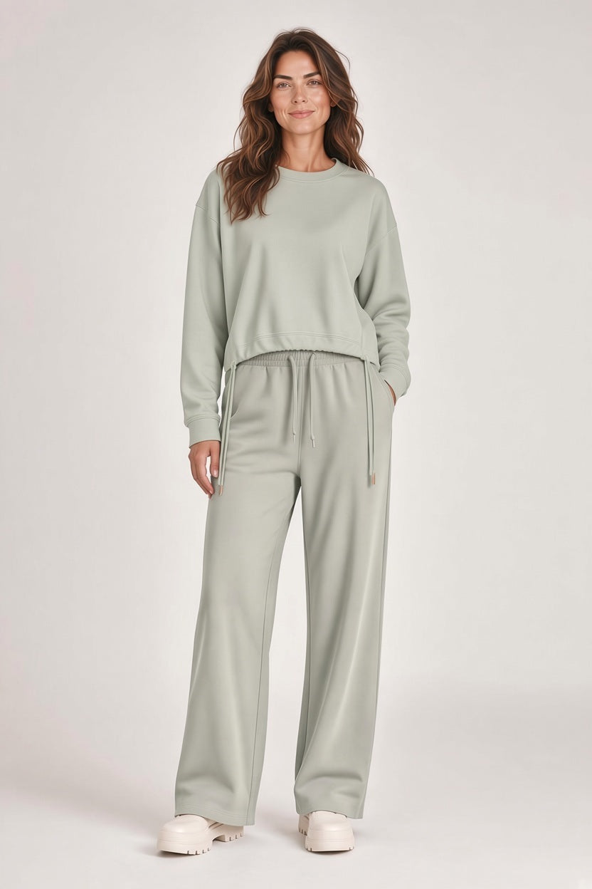 A woman wears the SAVE THE DUCK - AKEMI SWEATER IN RAINY BEIGE in a light green crewneck sweatshirt with matching wide-leg drawstring sweatpants and thick-soled shoes.