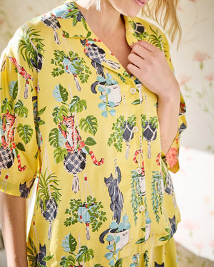 A woman stands barefoot indoors against a plain white wall and light wood floor, wearing the PRINTFRESH - Macrame Mischief Satin Wildest Dream Pajama Set in Sunshine by PRINTFRESH, featuring a loose-fitting eco satin yellow fabric with a colorful floral and cat print.