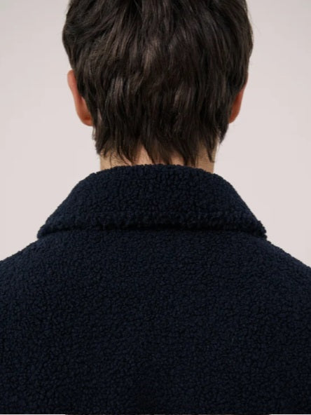 A young person with short dark hair wears the HARTFORD - Sherpa Utility Jacket in Navy by HARTFORD over a black shirt, looking directly at the camera against a plain light background. This warm sherpa layer brings both comfort and style to casual menswear.