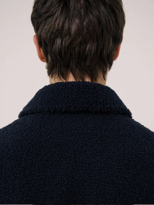 A young person with short dark hair wears the HARTFORD - Sherpa Utility Jacket in Navy by HARTFORD over a black shirt, looking directly at the camera against a plain light background. This warm sherpa layer brings both comfort and style to casual menswear.