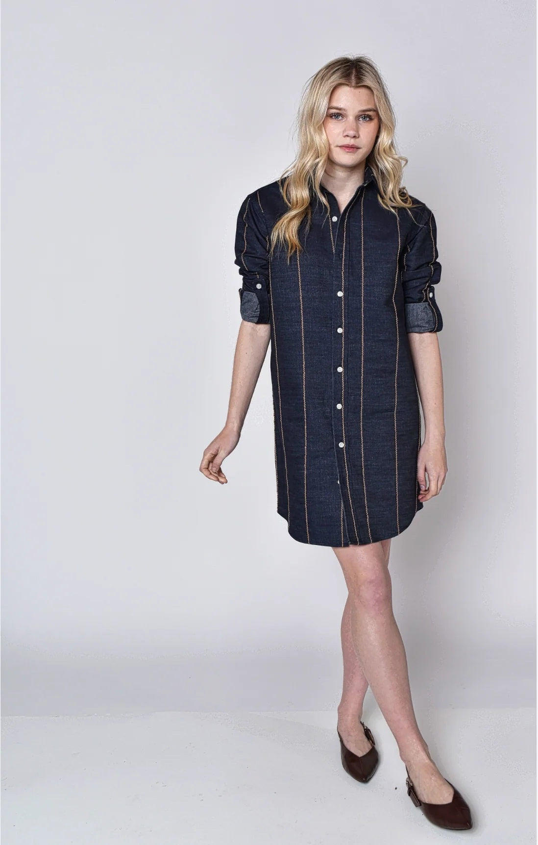 A person with long blonde hair wears the EMILY PHILLIPS Denim and Brown Stripe Girlfriend Mini Dress, styled with brown flats and rolled sleeves, standing against a plain light background.