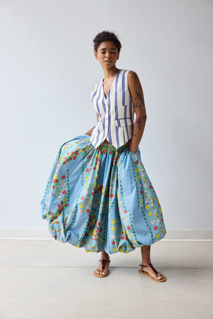 A person stands indoors wearing a white and blue striped vest and the HOPE FOR FLOWERS Bubble Hem Skirt in Chambray Floral by HOPE FOR FLOWERS. They have short curly hair and are wearing sandals.