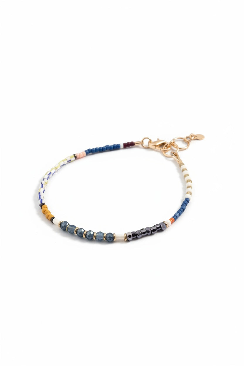 The ALICE RISE - SAPPHIRE GEMSTONE BRACELET by ALICE RISE, featuring multicolored beads and gold clasps, rests on a blue textured surface beside a white sand dollar—a striking example of elegant jewelry.